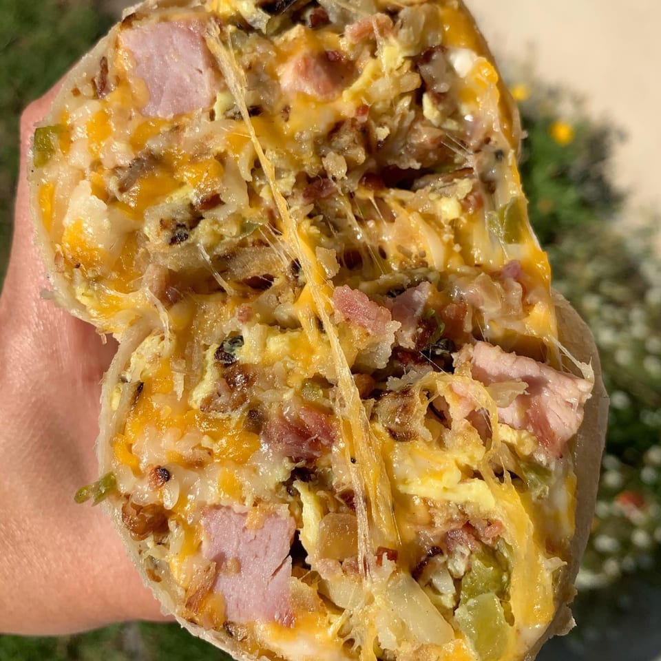 A hand holds a large, cut breakfast burrito filled with ham, cheese, eggs, and green peppers.