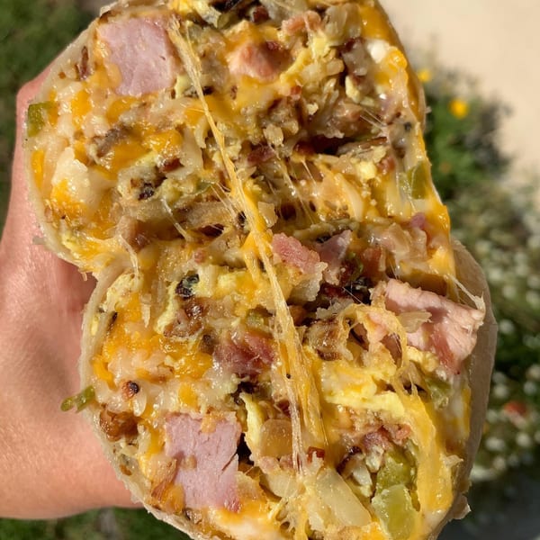 A hand holds a large breakfast burrito, cut in half, revealing melted cheese, ham, eggs, and potatoes.