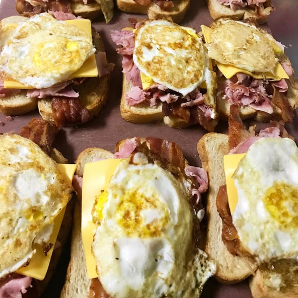Multiple open-faced breakfast sandwiches with fried eggs, cheese, ham, and bacon on toast.