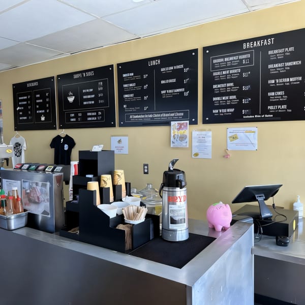 A restaurant counter with coffee, cups, and a pink piggy bank, with menu boards above.