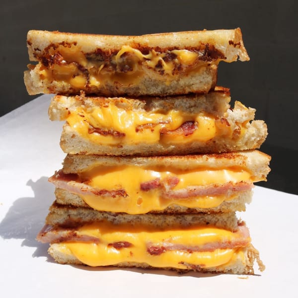 A stack of four grilled cheese sandwiches with melted cheese and bacon, on a white surface.