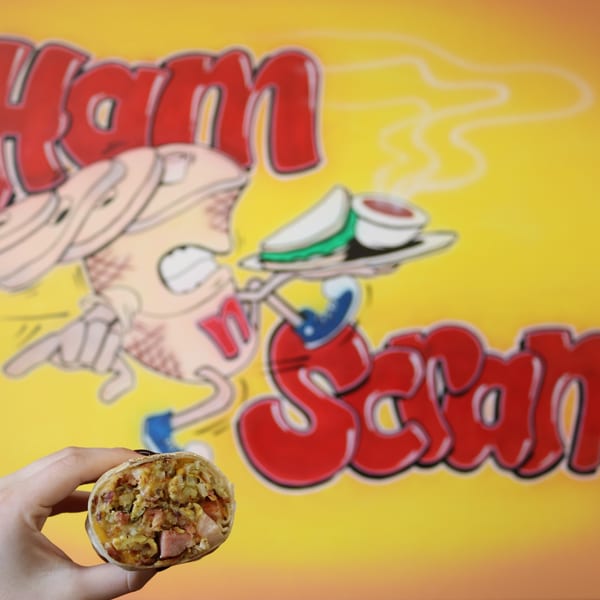 Hand holding a breakfast burrito filled with eggs, ham, and cheese, against a yellow Ham Scram cartoon logo.